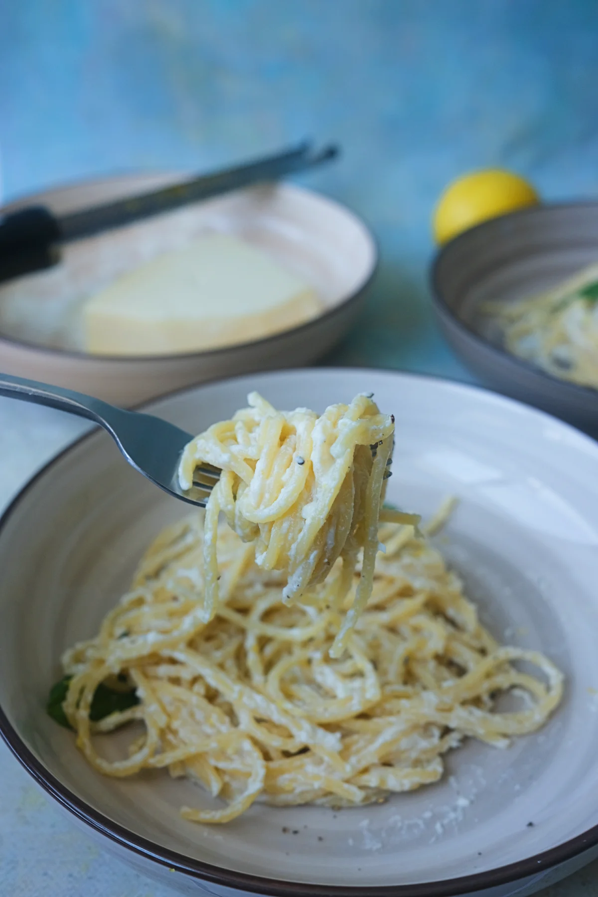 A fork holding a bite of creamy lemon ricotta spaghetti above a bowl of pasta, with a bowl of ricotta cheese, a lemon, and a microplane grater blurred in the background.