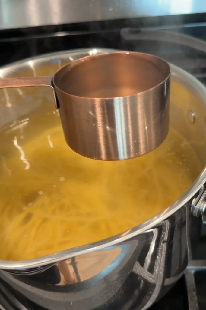 A measuring cup filled with reserved pasta water held above a pot of boiling spaghetti on the stovetop.