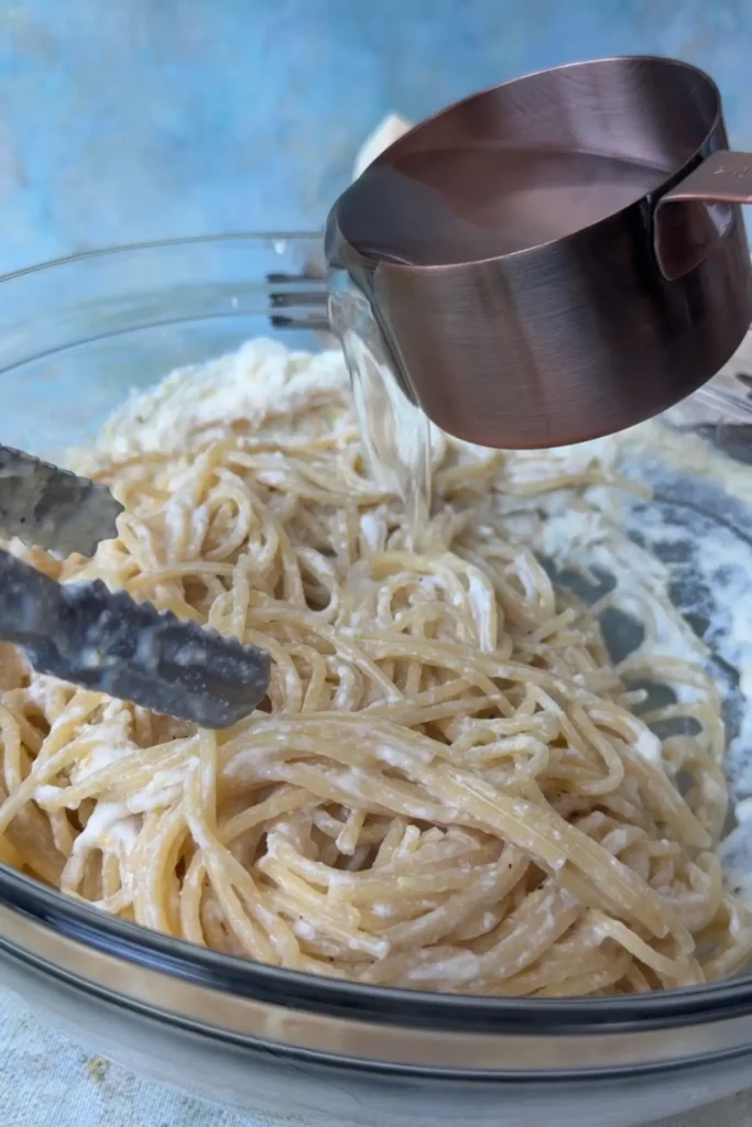 Reserved pasta water being poured into spaghetti coated with ricotta sauce while mixing with kitchen tongs in a glass bowl.