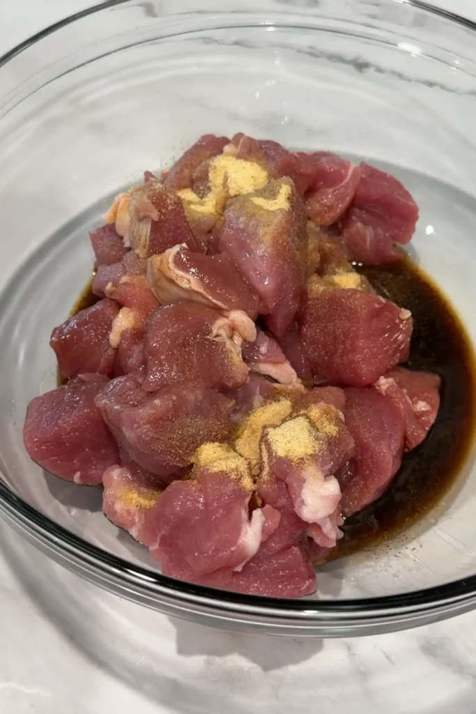 Cubed pork loin in a glass bowl with Worcestershire sauce, soy sauce, liquid seasoning, garlic powder, and black pepper before marinating.