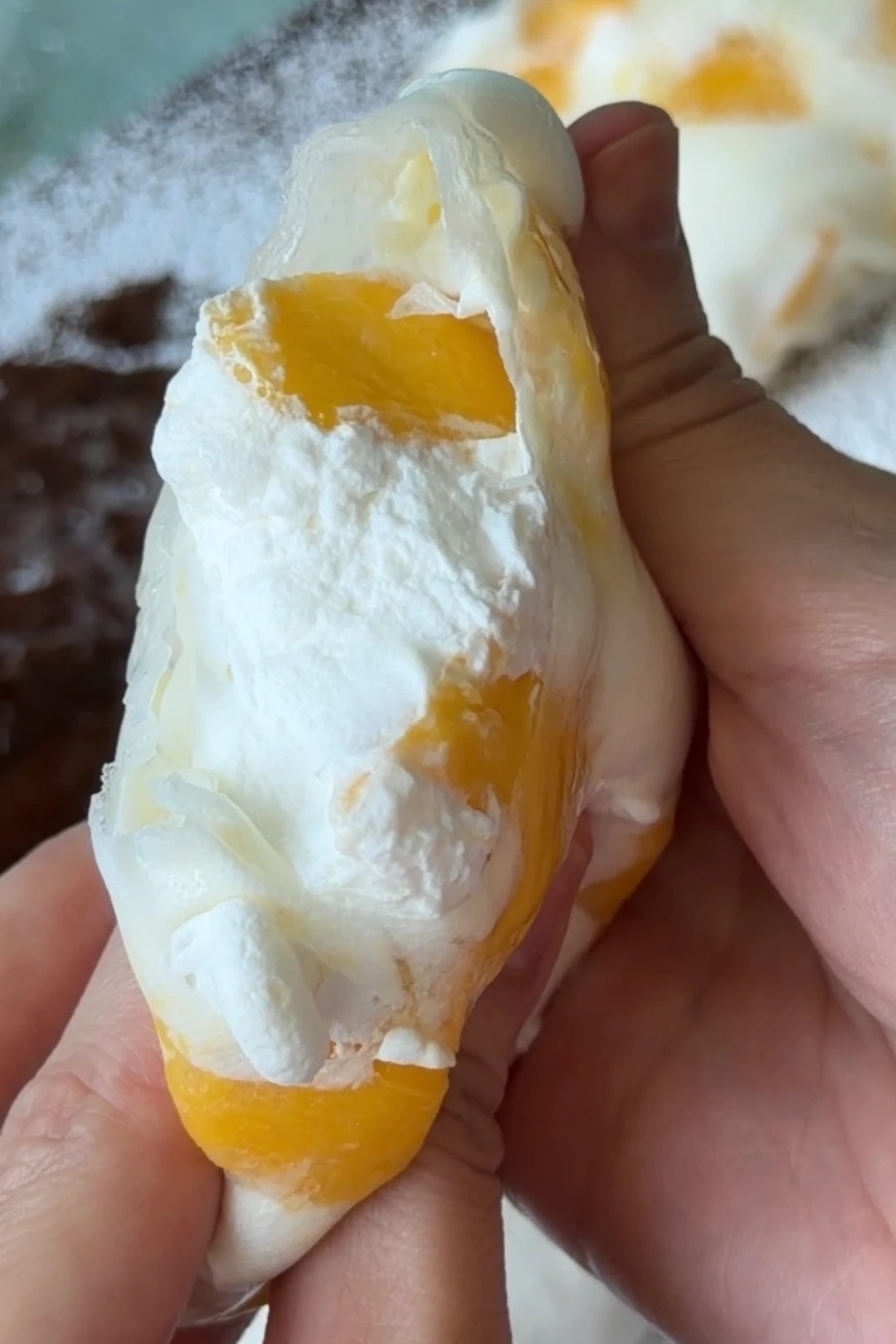 A hand holding a rice paper mango mochi with a bite taken out, revealing the whipped mango cream filling and fresh mango pieces.
