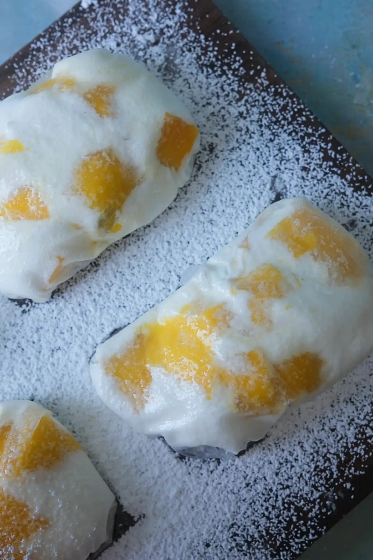 Overhead view of homemade rice paper mango mochi dusted with a potato starch and powdered sugar mixture on a dark serving board.