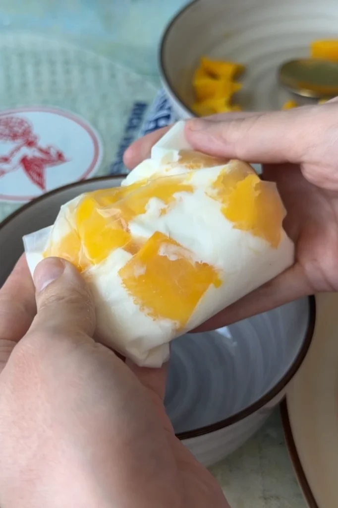 A freshly wrapped rice paper mango mochi held in both hands, showing fresh mango pieces through the translucent rice paper.