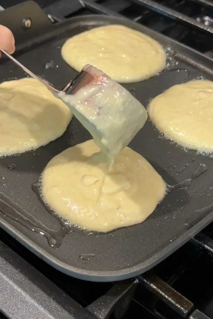Thick pancake batter being scooped onto a buttered nonstick griddle using a batter dispenser.