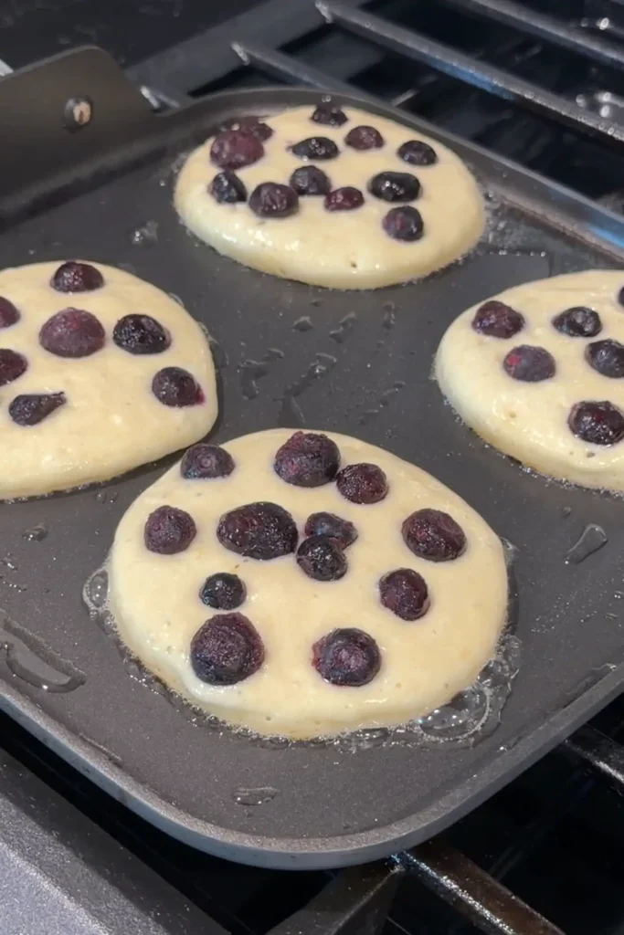 Frozen blueberries scattered over uncooked lemon ricotta pancake batter on a hot griddle.