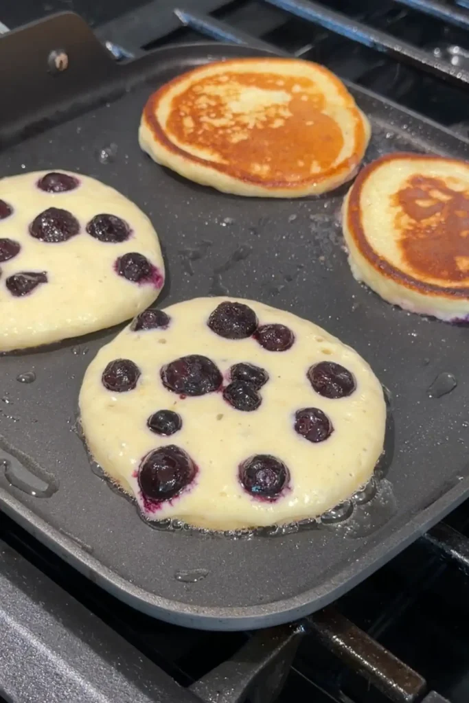 Lemon blueberry ricotta pancakes cooking on a griddle with golden brown pancakes after being flipped.