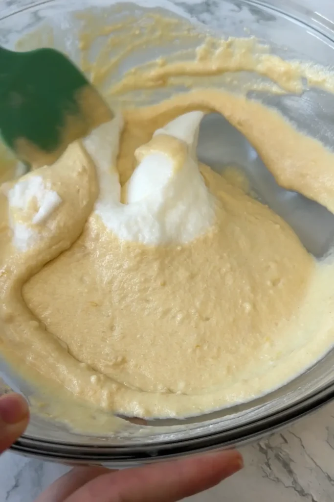 Beaten egg whites being gently folded into the pancake batter with a green silicone spatula.