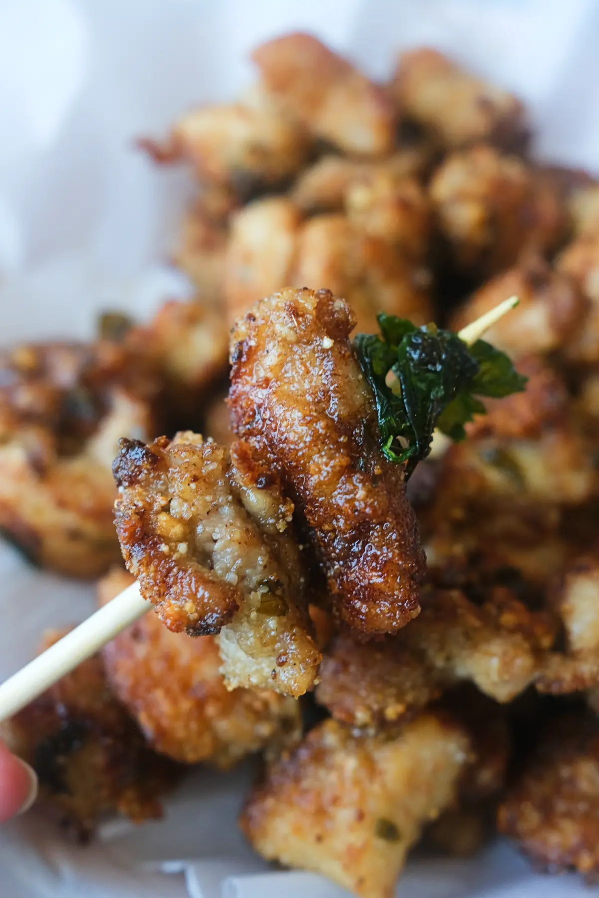 A skewer holding two pieces of Taiwanese popcorn chicken with crispy basil in front of a tray of chicken.