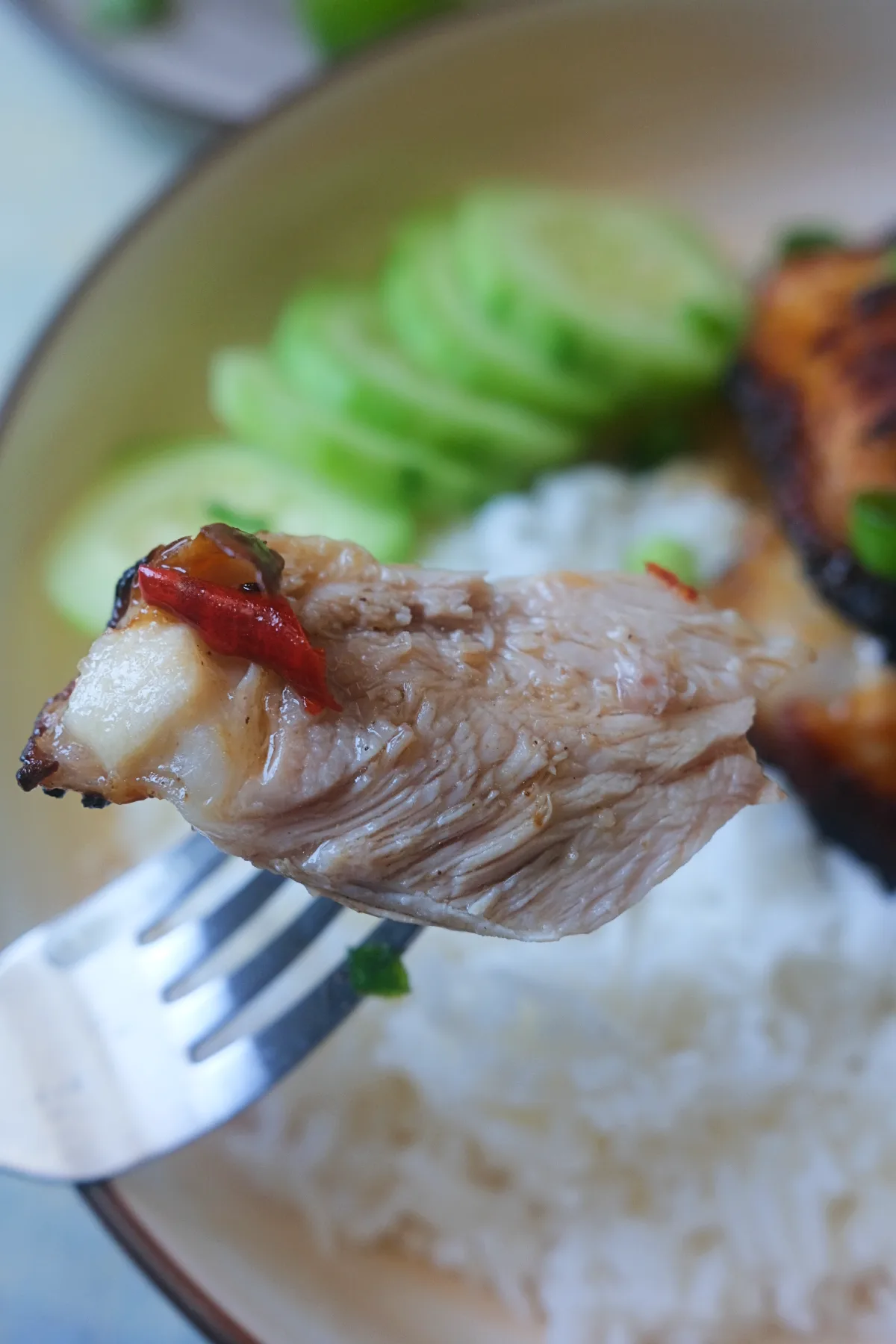 Fork holding a tender piece of Vietnamese lemongrass chicken showing the juicy inside with crispy edges, served with rice, cucumber, herbs, and fresh chilies.