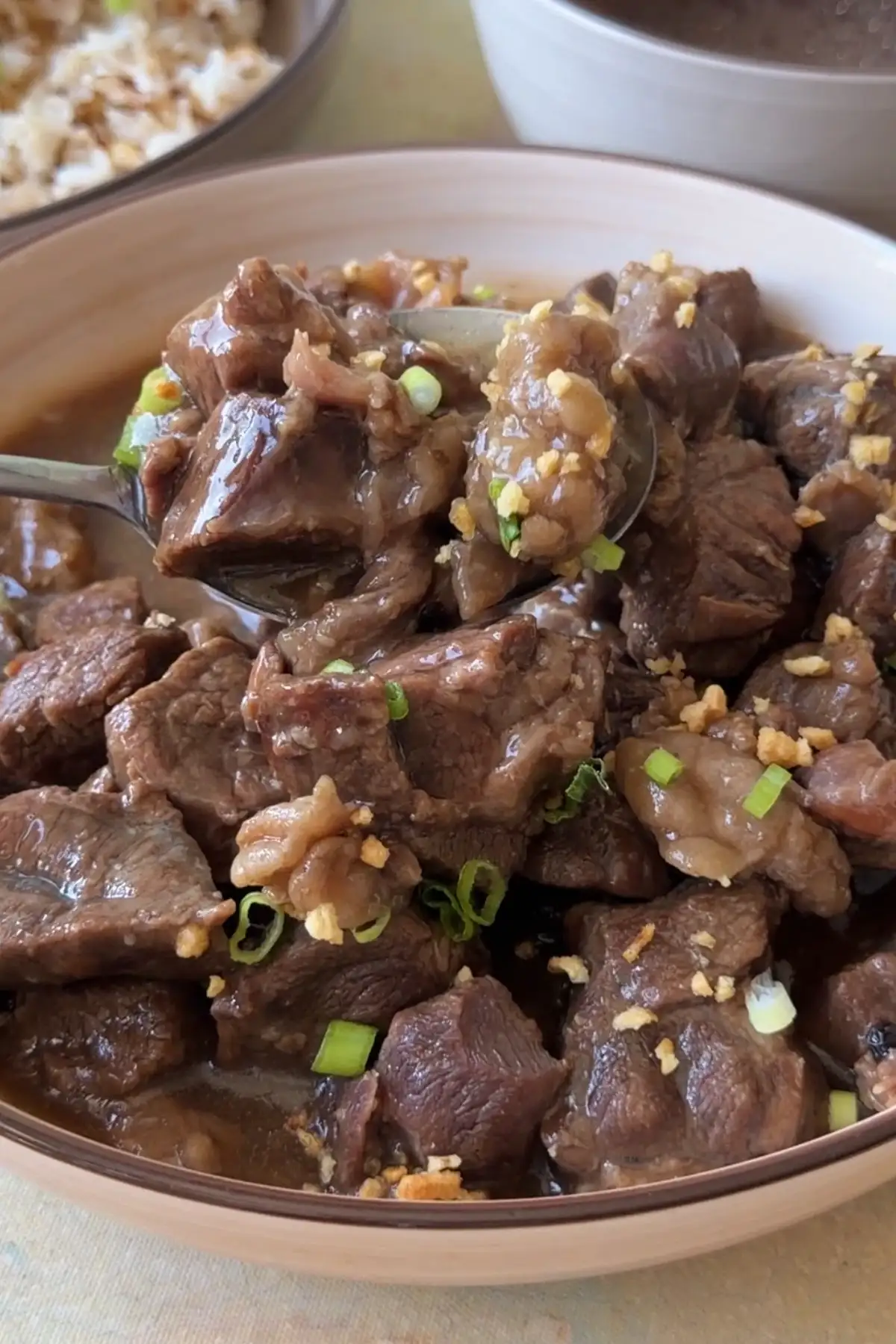 Spoon lifting tender pieces of Filipino beef pares topped with fried garlic and green onions.