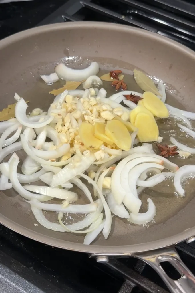Sliced onions, chopped garlic, ginger, bay leaves, star anise, and peppercorns added to a pan with oil.