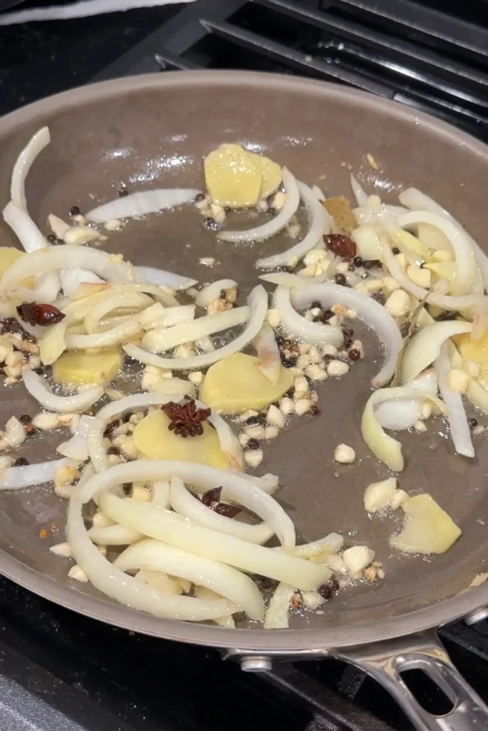 Onions, garlic, ginger, star anise, bay leaves, and peppercorns sautéing in a pan.