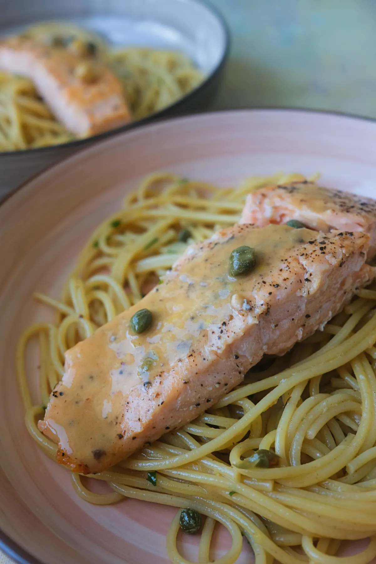 Salmon piccata with buttery lemon caper sauce served over spaghetti on a pink plate.