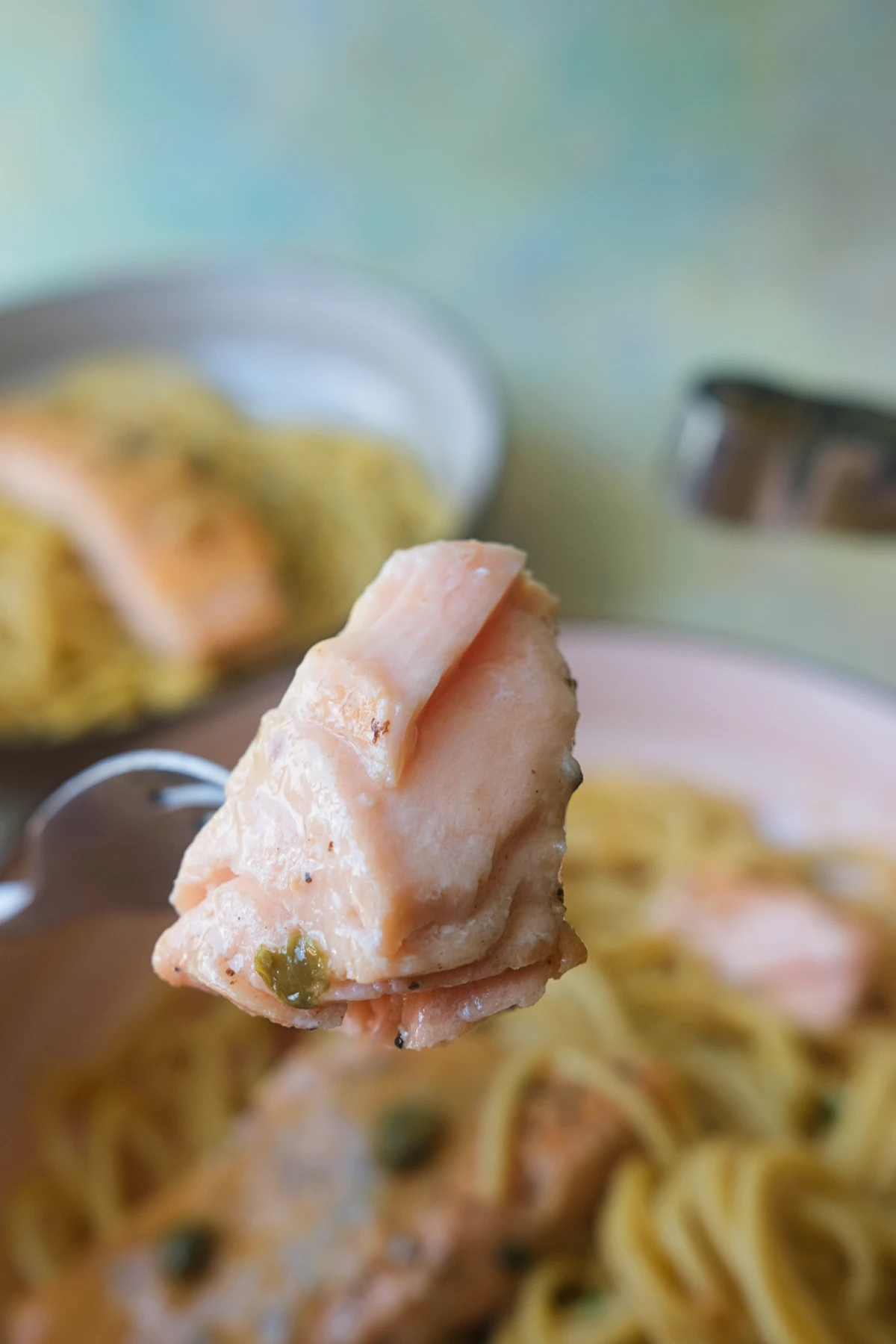 Fork holding a tender piece of salmon piccata over a plate of spaghetti.