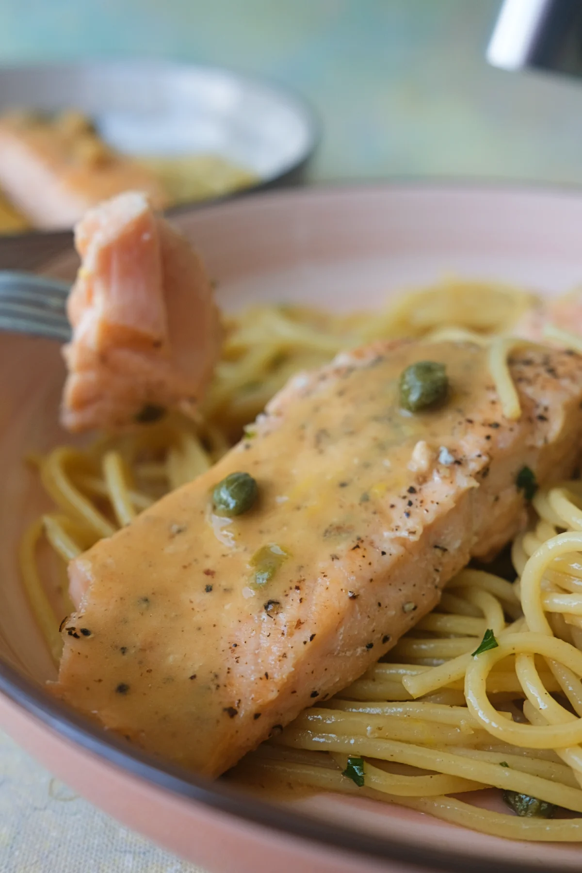 Salmon fillet topped with creamy lemon caper sauce over spaghetti with a bite lifted on a fork.