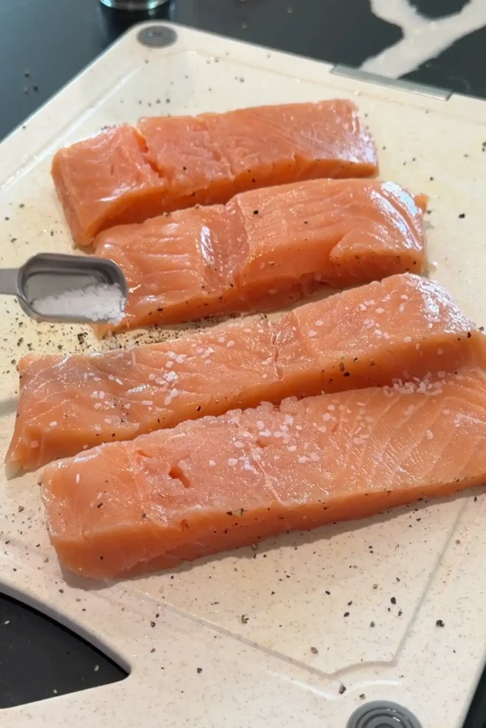 Raw salmon fillets seasoned with salt and black pepper on a cutting board.
