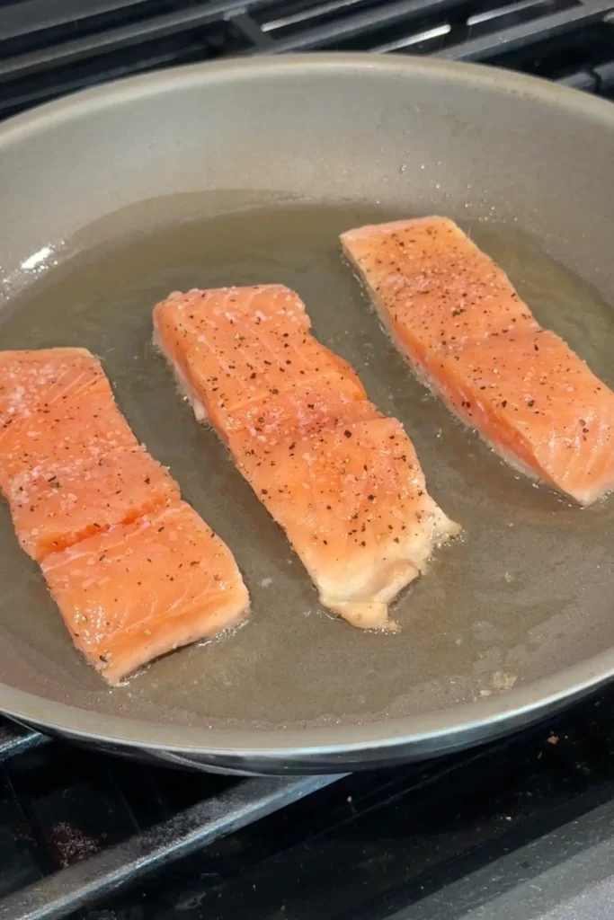 Seasoned salmon fillets searing in oil in a large skillet.