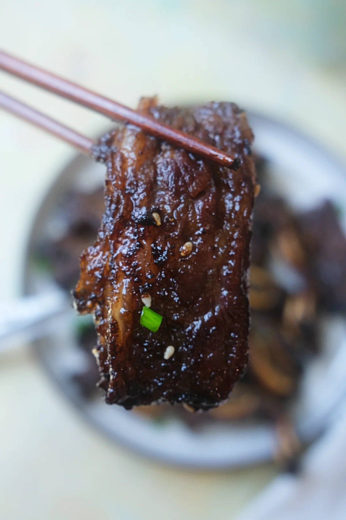 Close-up of chopsticks holding a caramelized piece of Korean beef short rib with green onions and sesame seeds.
