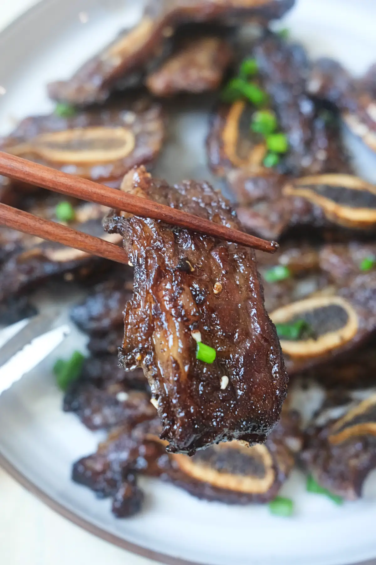 Chopsticks holding a tender piece of air fryer Korean short rib over a plate of cooked ribs.