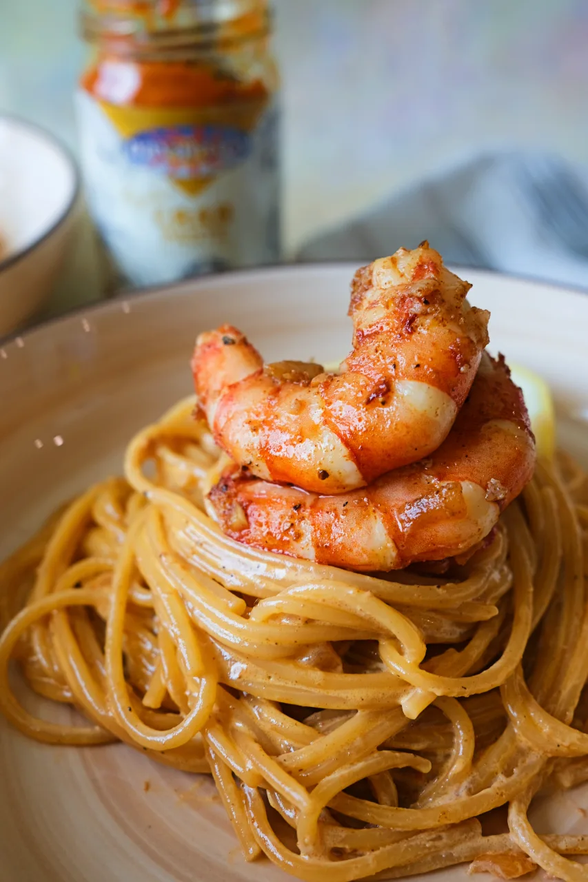 Close-up of creamy aligue pasta topped with sautéed shrimp, with a jar of crab paste in the background.