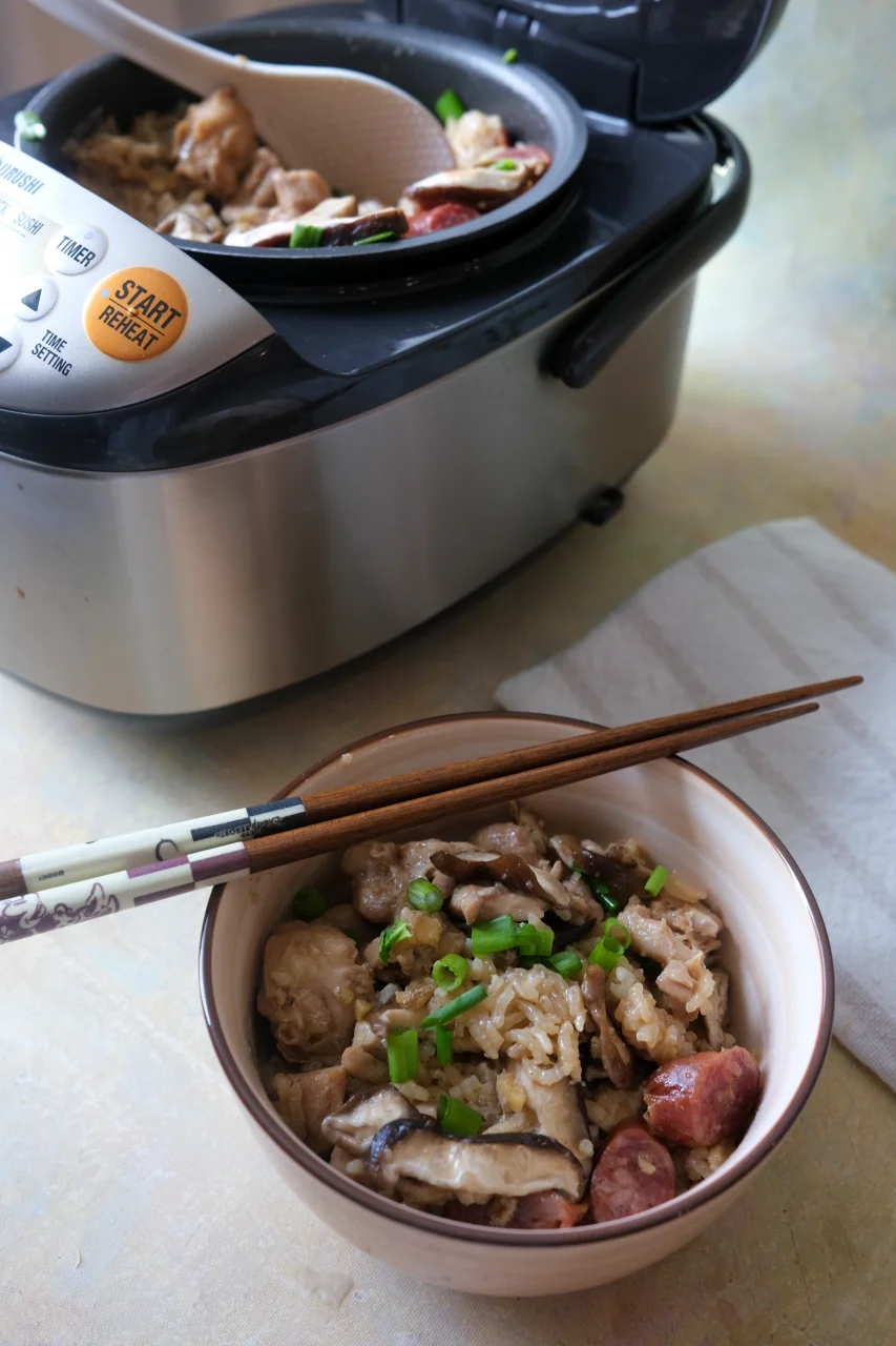 Bowl of claypot chicken rice beside an open rice cooker filled with chicken, sausage, mushrooms, and rice.