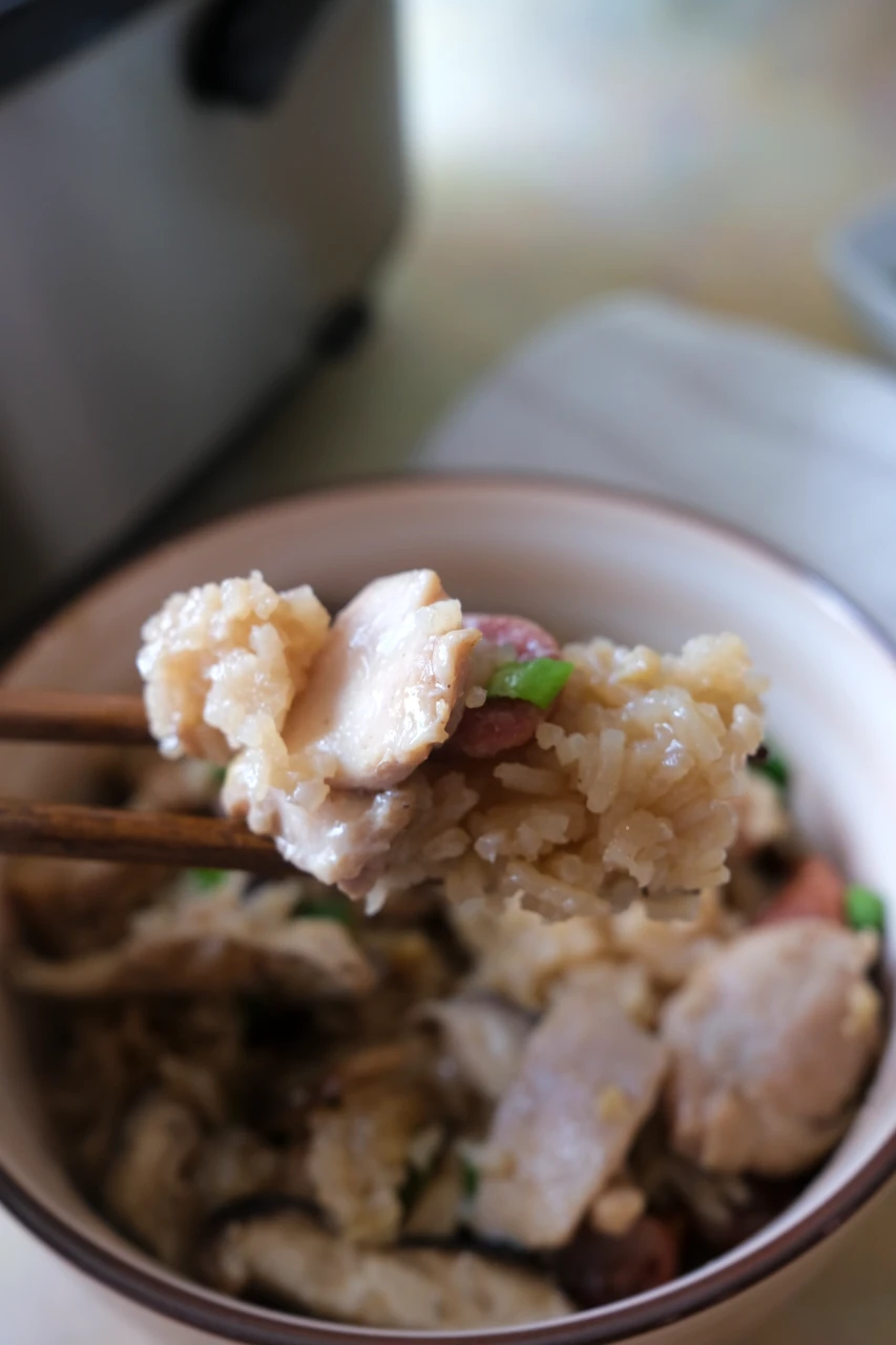 Chopsticks holding a bite of chicken and seasoned rice over a bowl of rice cooker claypot chicken rice.