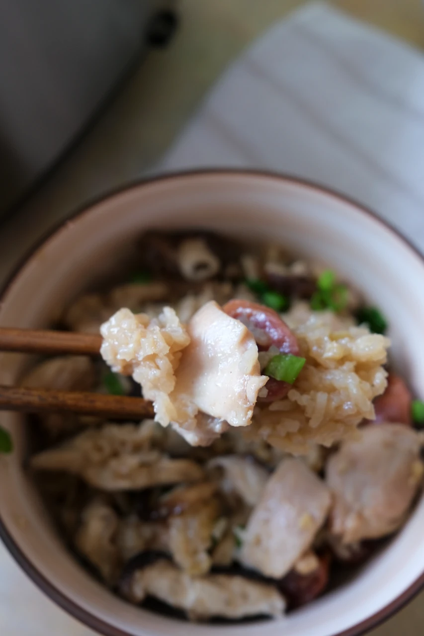 Chopsticks holding chicken and rice above a bowl of claypot chicken rice with Chinese sausage and mushrooms.