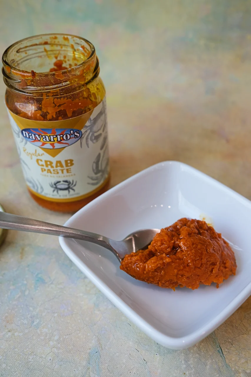 Jar of crab paste beside a spoonful of reddish-orange aligue in a white dish.