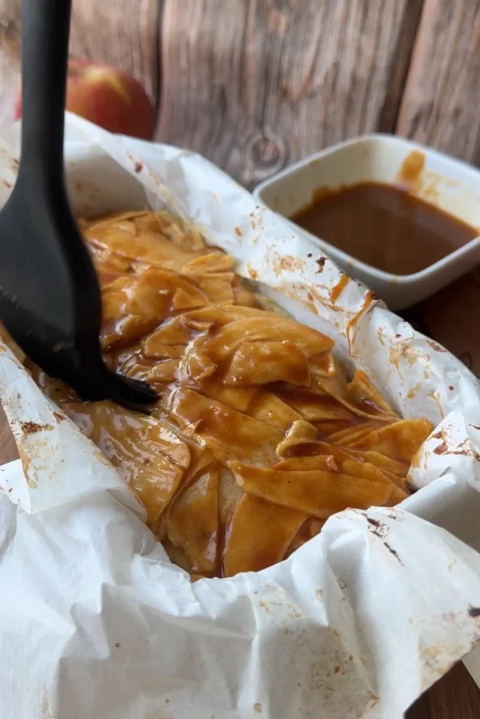 Brushing caramel sauce over the top of warm invisible apple cake in a loaf pan.
