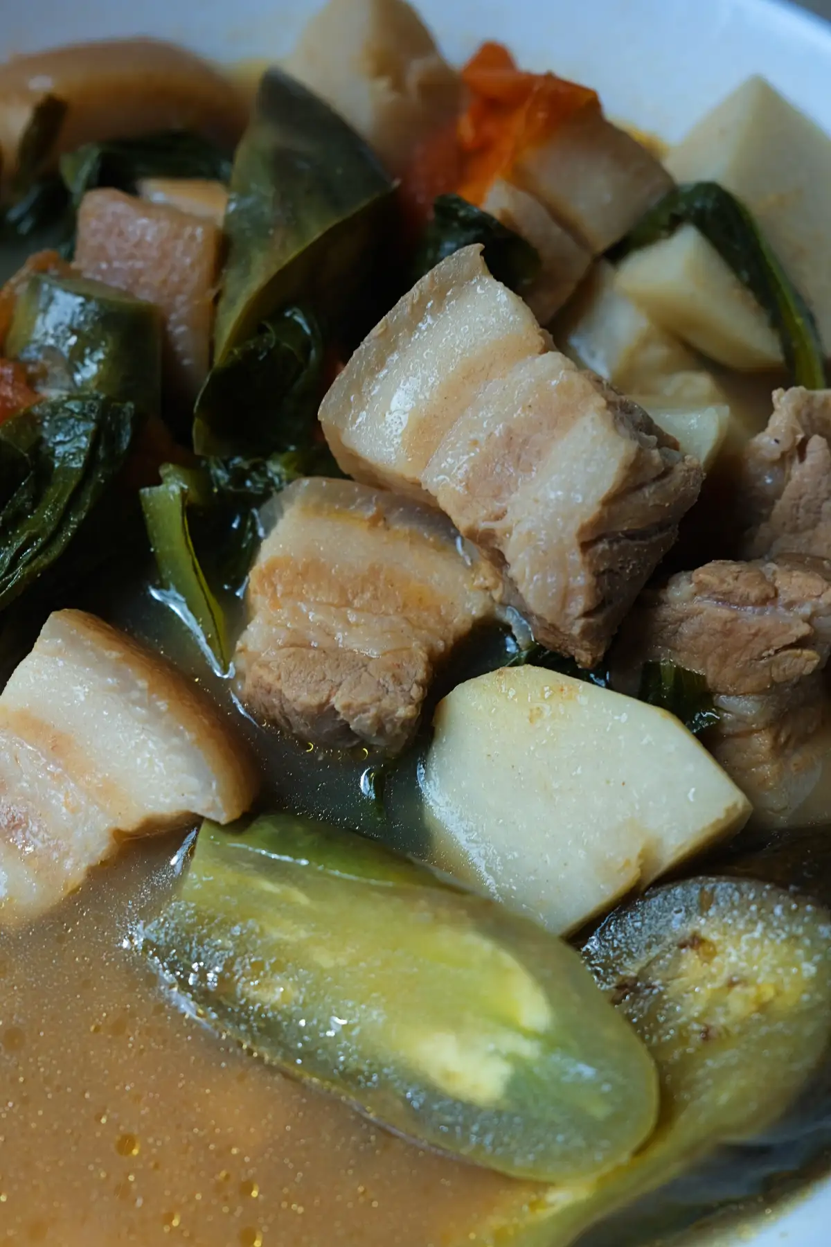 Close-up of pork sinigang with tender pork belly, eggplant, taro, leafy greens, and vegetables in a sour broth.