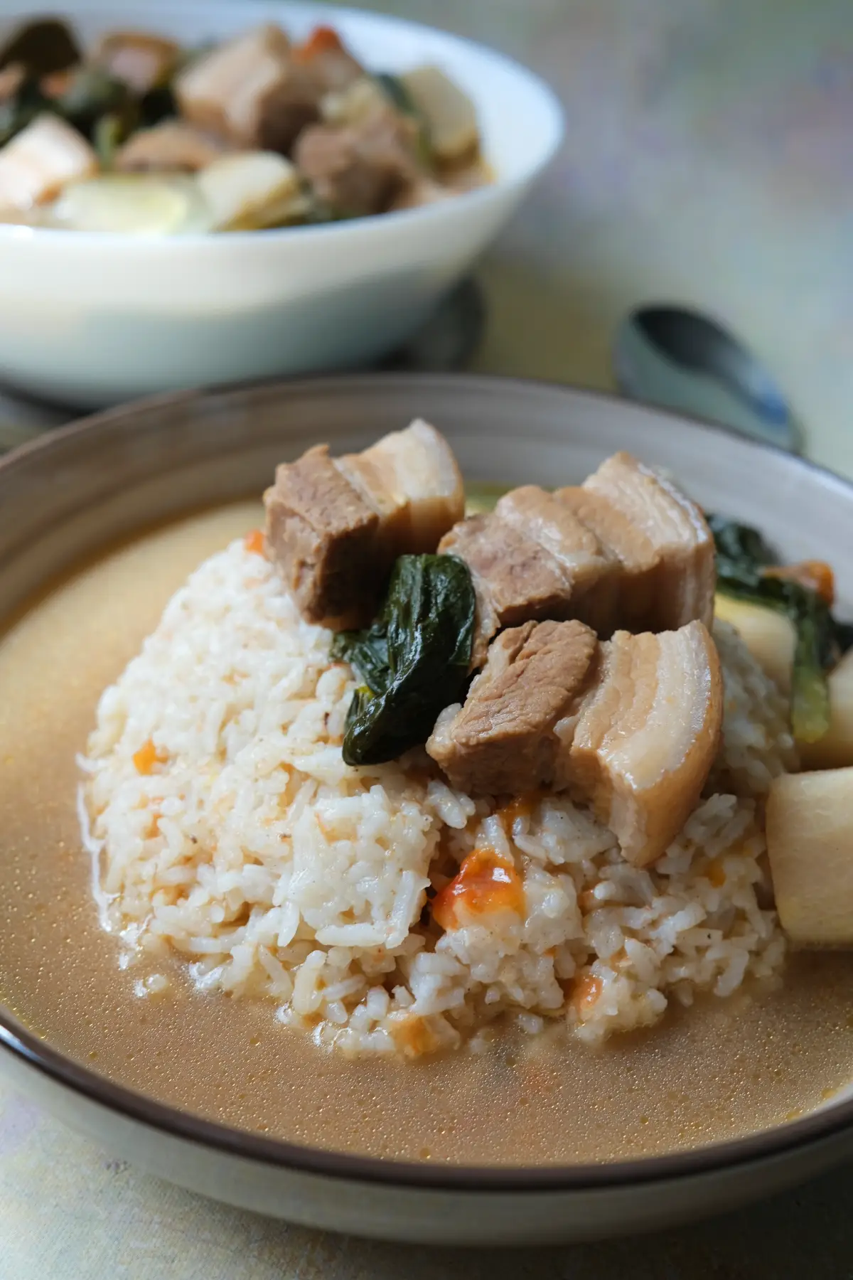 Bowl of pork sinigang with white rice topped with pork belly and leafy greens, surrounded by vegetables and broth.