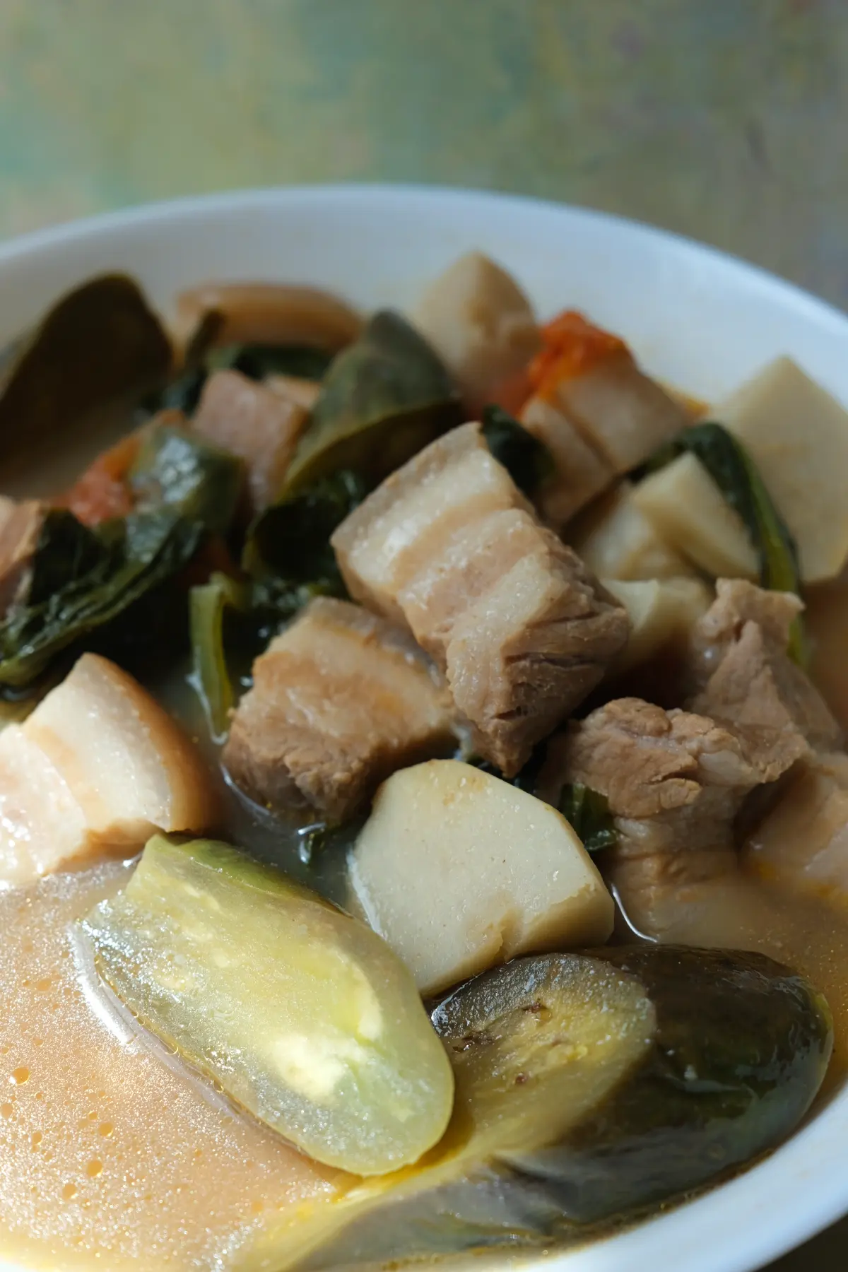 Close-up of Filipino pork sinigang with thick pieces of pork belly, eggplant, taro, and leafy greens in sour broth.
