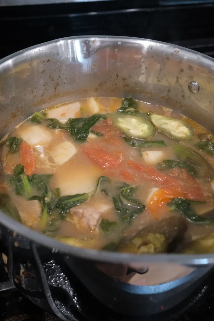 Pork sinigang simmering in a pot with pork belly, tomatoes, eggplant, taro, and leafy greens.