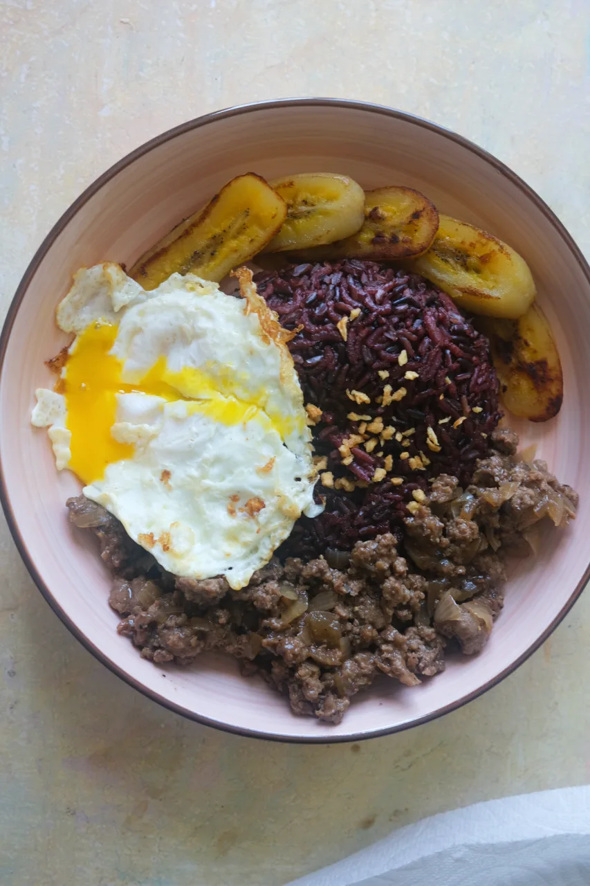 Overhead view of Filipino Arroz a la Cubana with black rice, ground beef, fried saba bananas, and a runny fried egg.