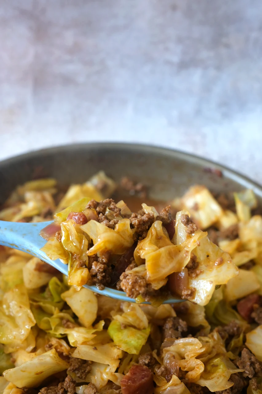 Blue serving spoon lifting cooked cabbage, ground beef, sausage, and onion from the skillet.