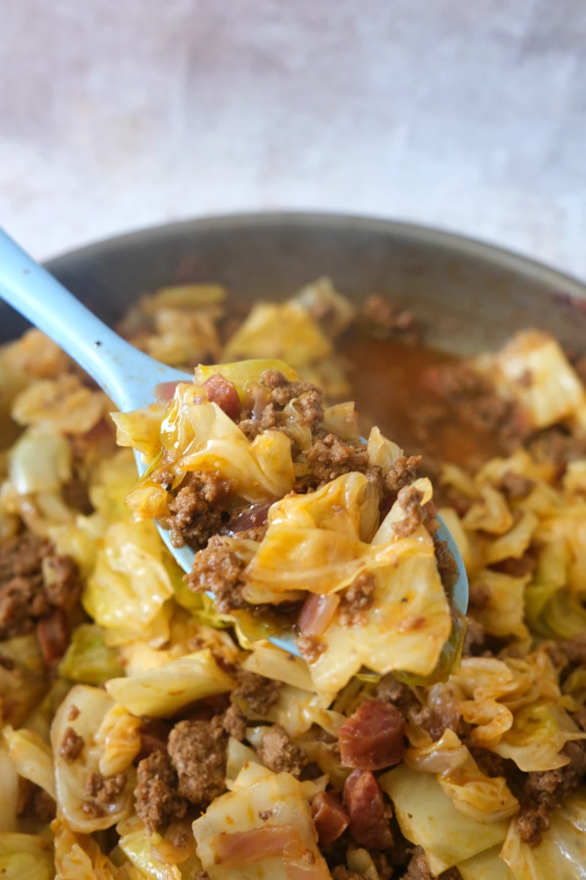 Blue serving spoon lifting cooked cabbage, ground beef, sausage, and onion from the skillet.