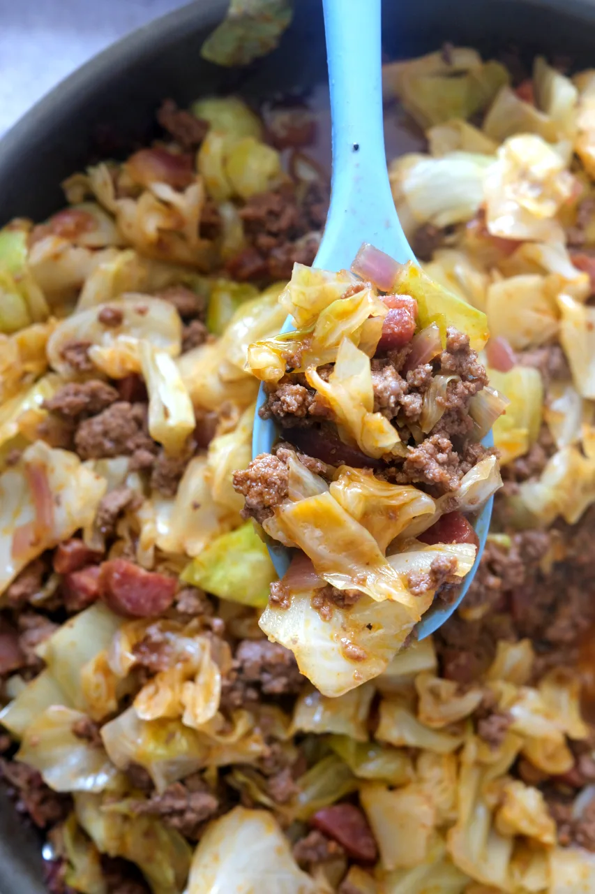 Overhead view of a blue spoon holding ground beef and cabbage above the skillet.