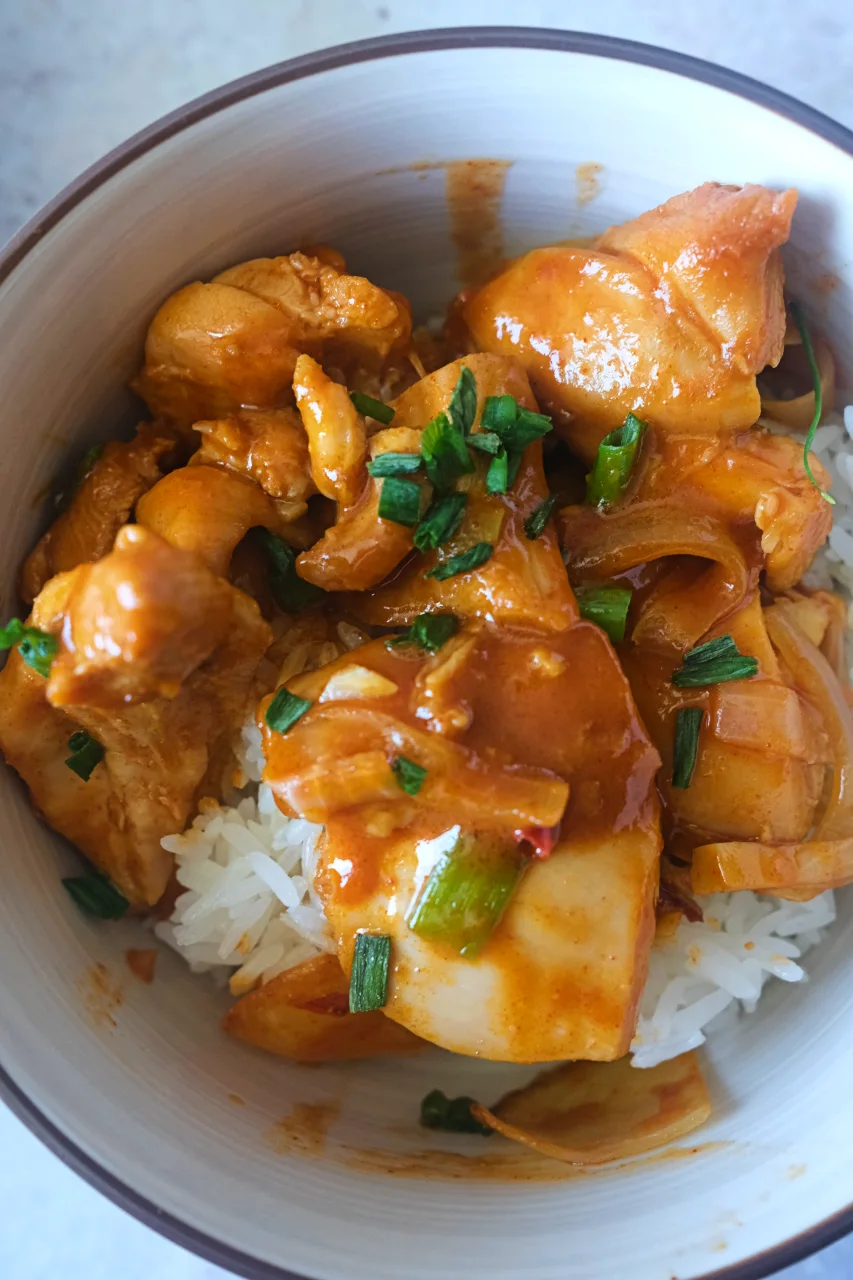 Overhead view of chicken thigh pieces coated in gochujang sauce with onions and green onion garnish.