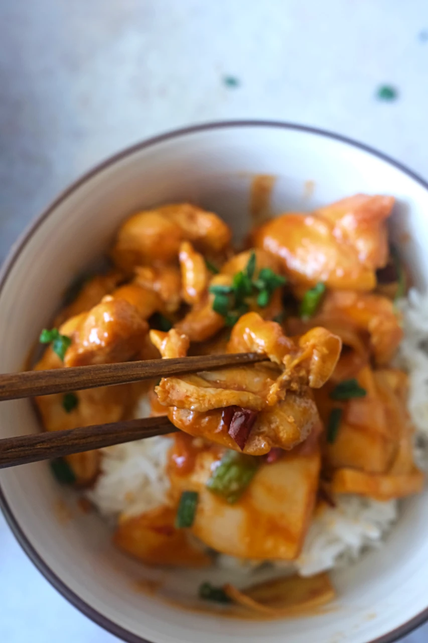 Chopsticks holding a piece of gochujang chicken above a bowl of chicken and white rice.