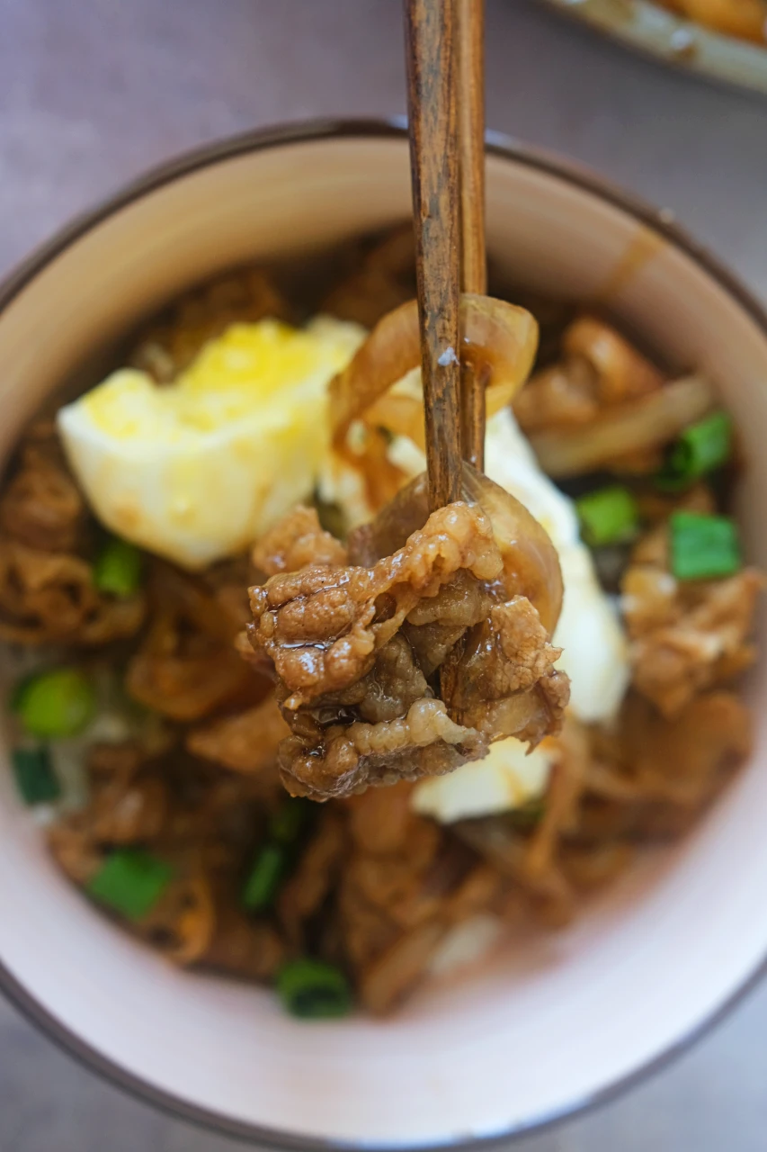 Chopsticks lifting thinly sliced beef and onion above a gyudon bowl topped with egg and green onions.