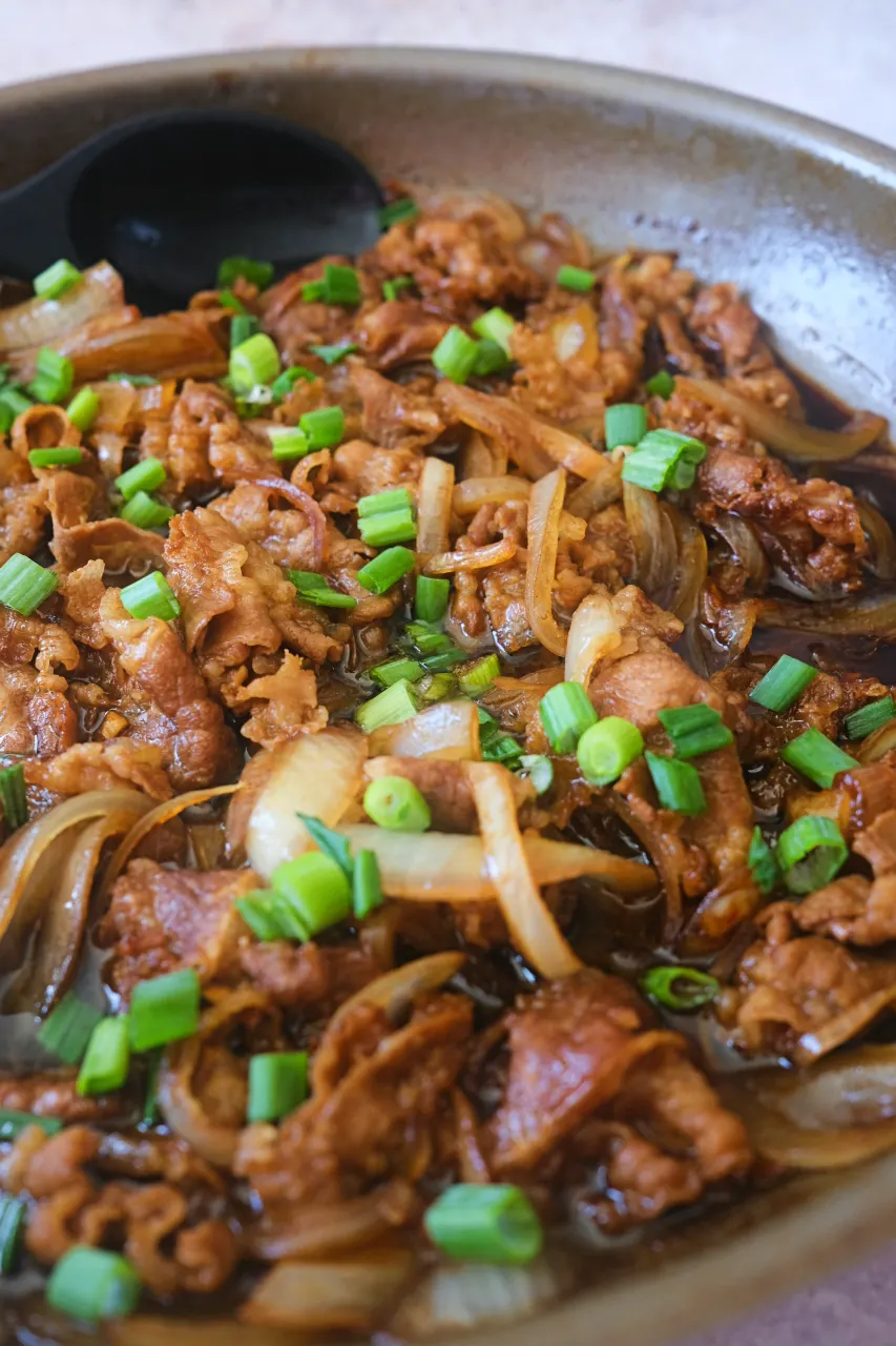 Beef gyudon with sliced onions and chopped green onions in a skillet of brown sauce.