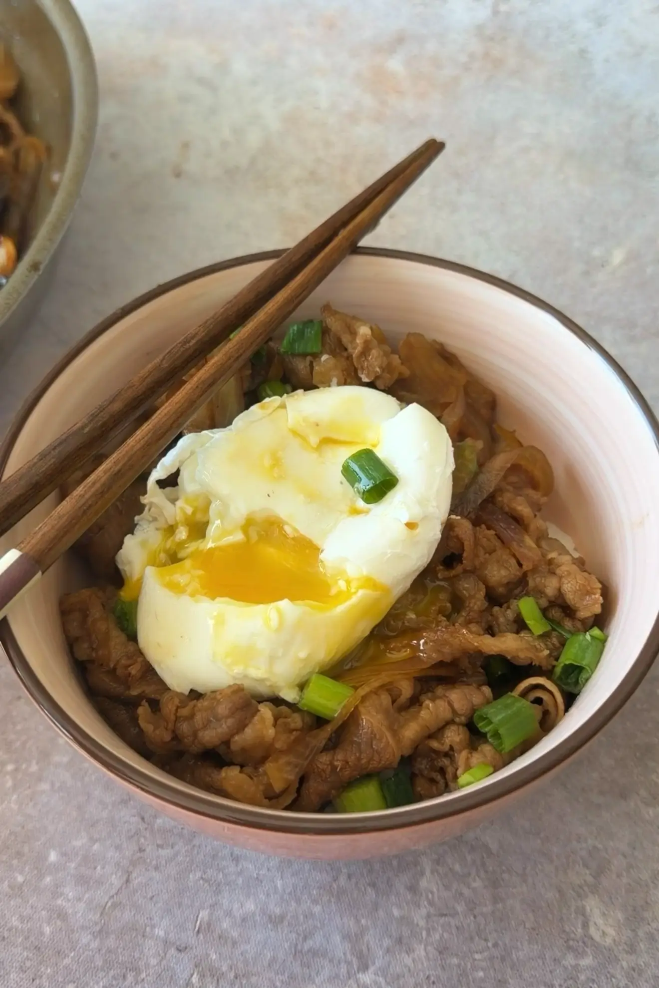 Japanese beef rice bowl topped with a split poached egg, runny yolk, and green onions, with chopsticks resting across the bowl.