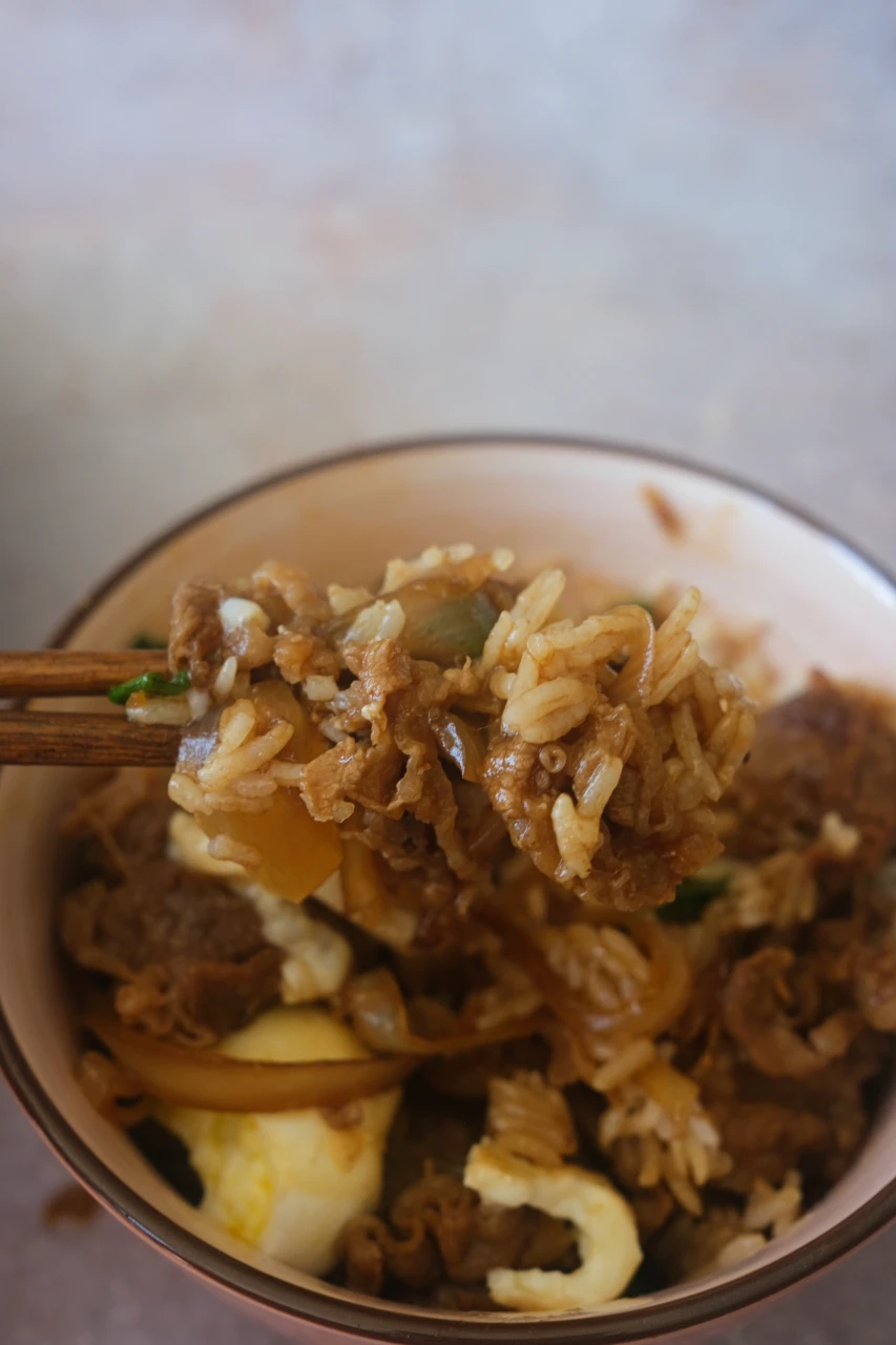 Chopsticks holding a bite of beef, onion, and sauce-coated rice above a gyudon bowl.