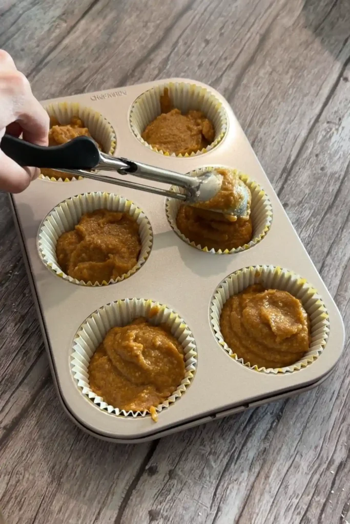 Pumpkin muffin batter being portioned into six lined muffin cups with a scoop.