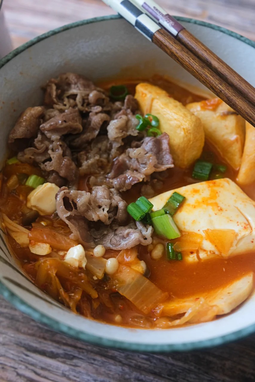 Bowl of Korean kimchi jjigae topped with thinly sliced beef, tofu, mushrooms, and green onions.