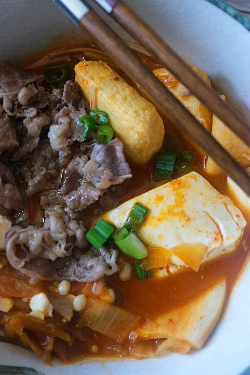 Close-up of spicy kimchi stew with sliced beef, soft tofu, hot pot balls, and green onions.